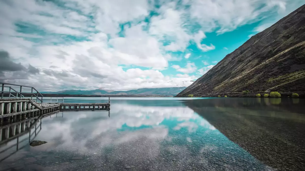 Clear lake with mountain slopes, wooden pier, and clouds reflected in the water at Lake Ohau. Main Image